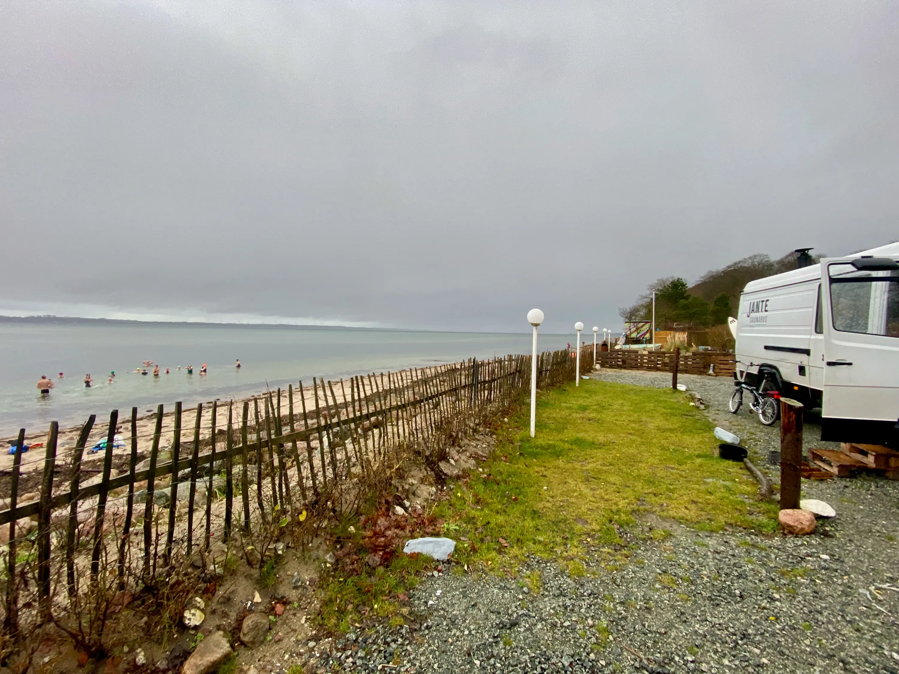 Schwimmerinnen und Schwimmer im flachen, grauen Wasser einer Bucht; rechts steht der Saunabus hinter einem Strandzaun.