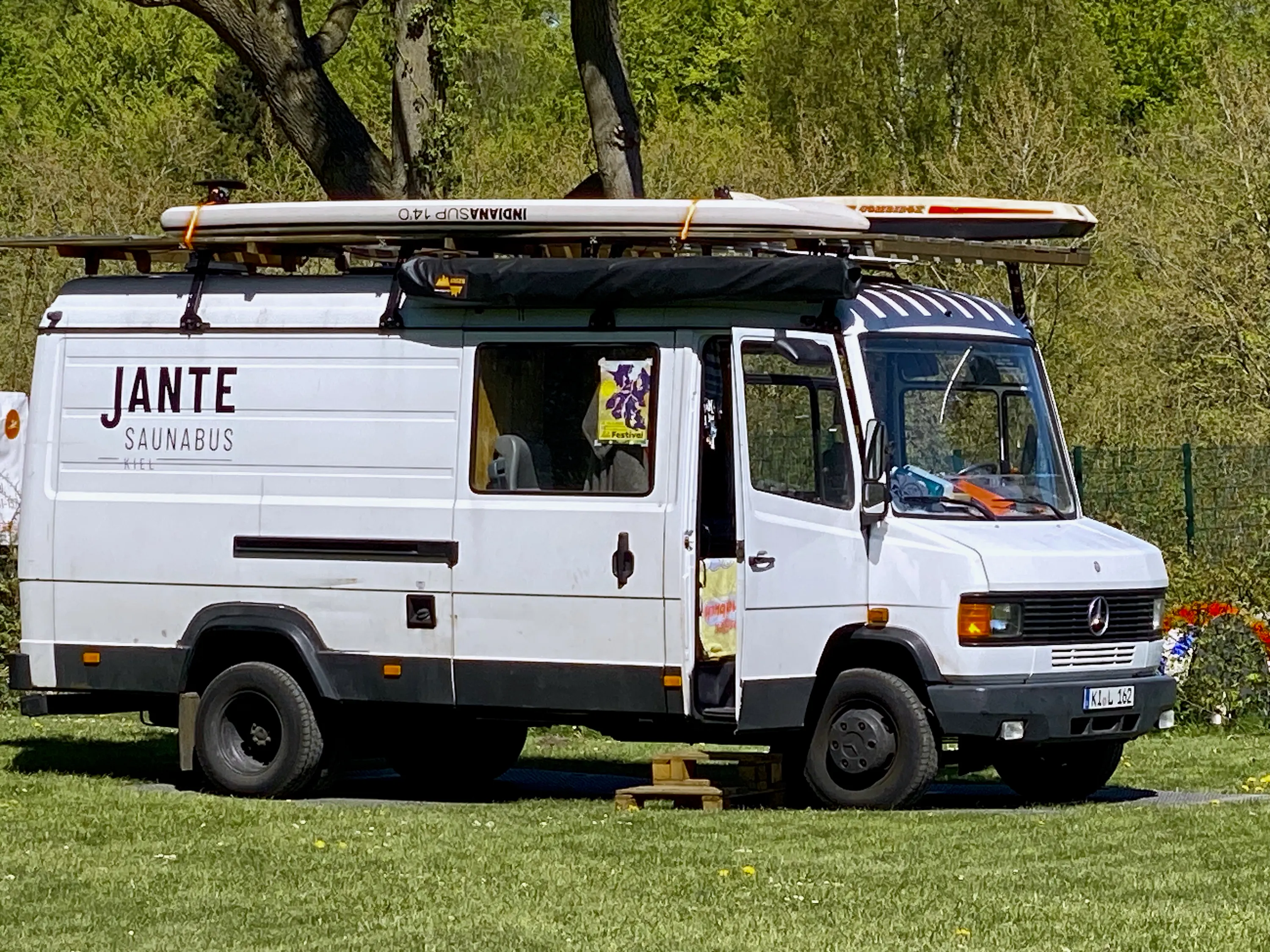 Der weiße Saunabus mit zwei Stand-up-Paddleboards auf dem Dach parkt auf einer sonnigen Wiese.
