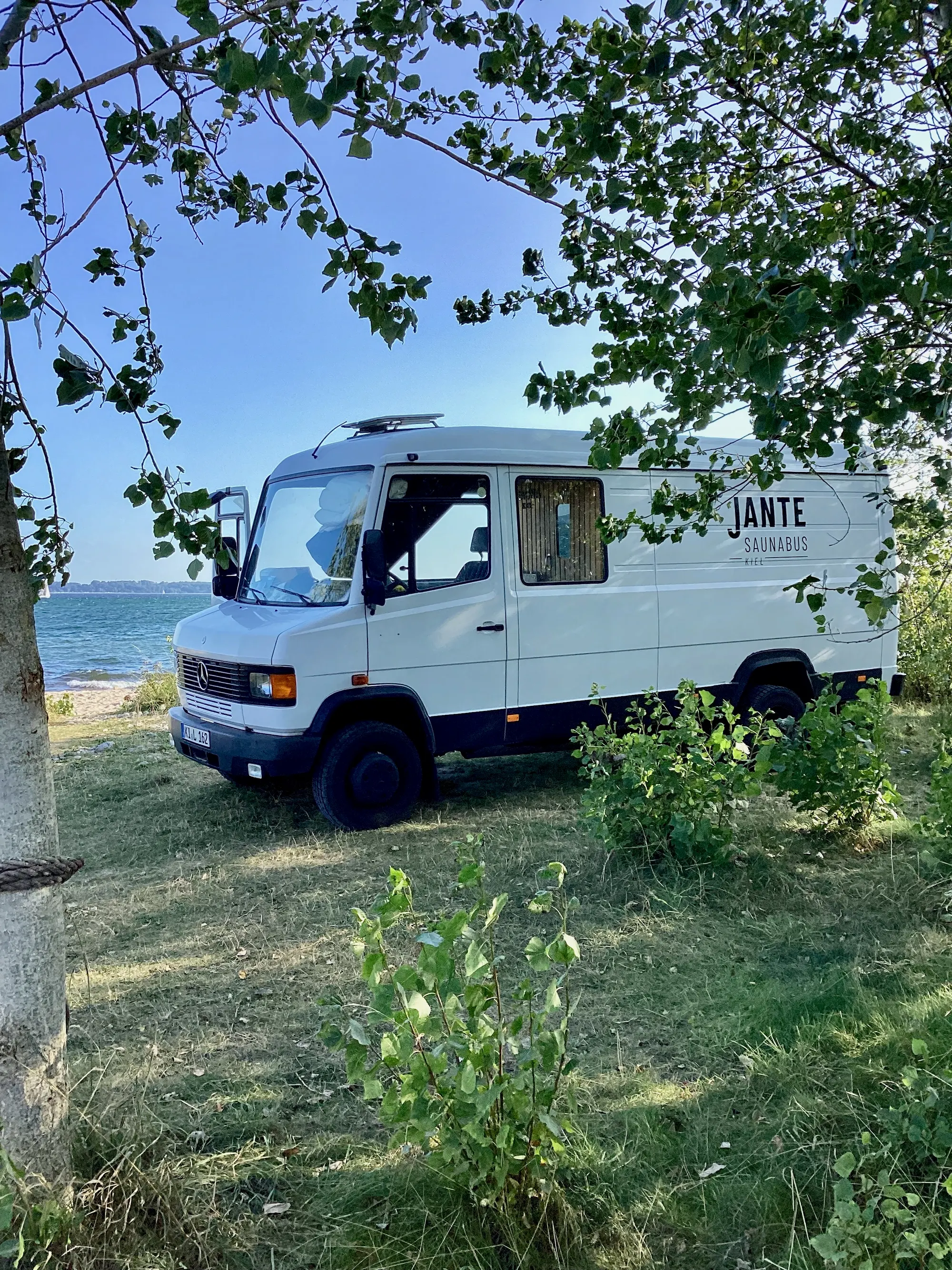 Der Saunabus auf einer Wiese direkt am Ufer, von Blättern umrahmt, dahinter das Meer.