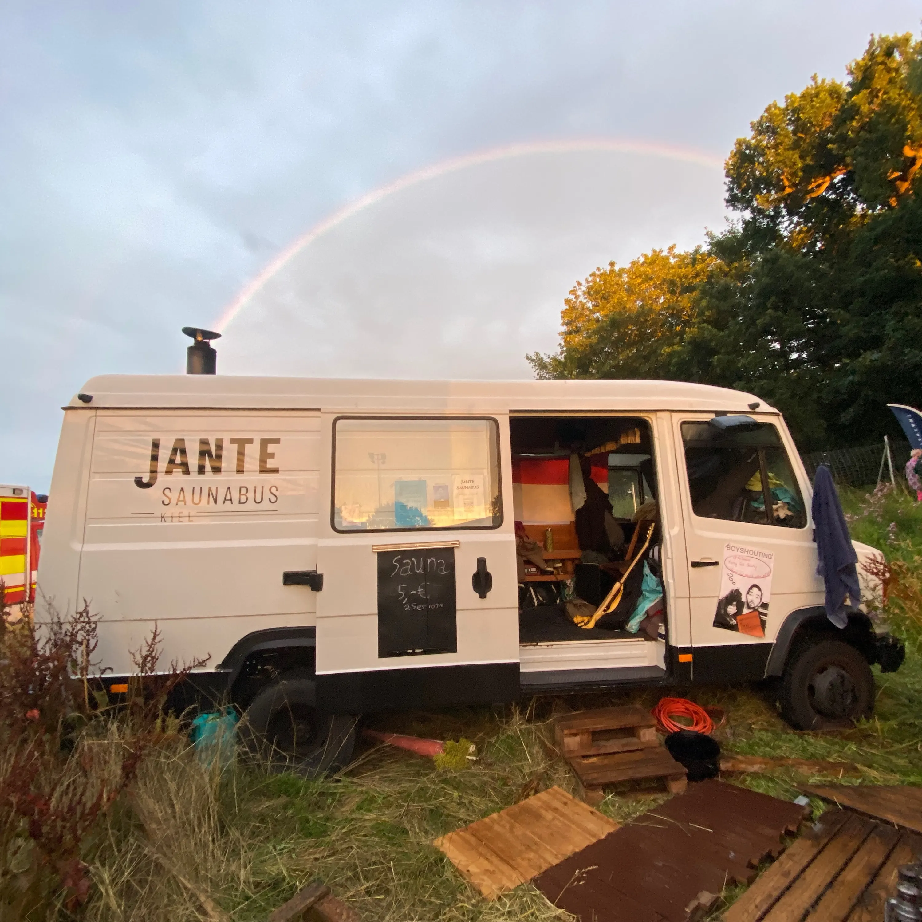 Ein Regenbogen über dem Saunabus, der mit offener Tür auf einer Wiese steht, an der Seite eine Kreidetafel.