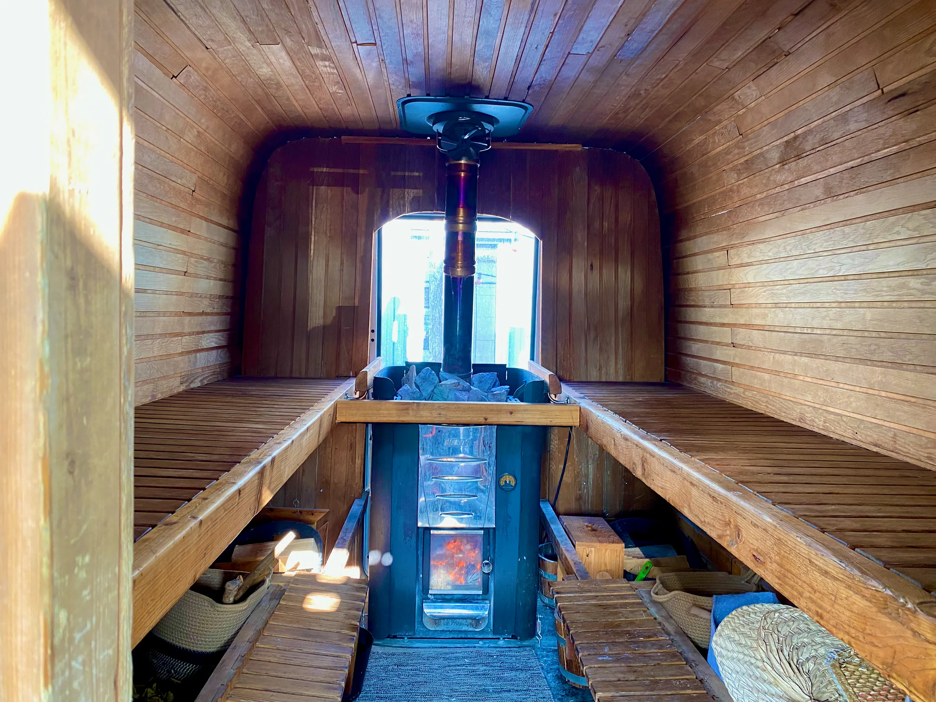 Innenraum der Sauna: Holzbänke links und rechts, dazwischen der Ofen unter einem Fenster.