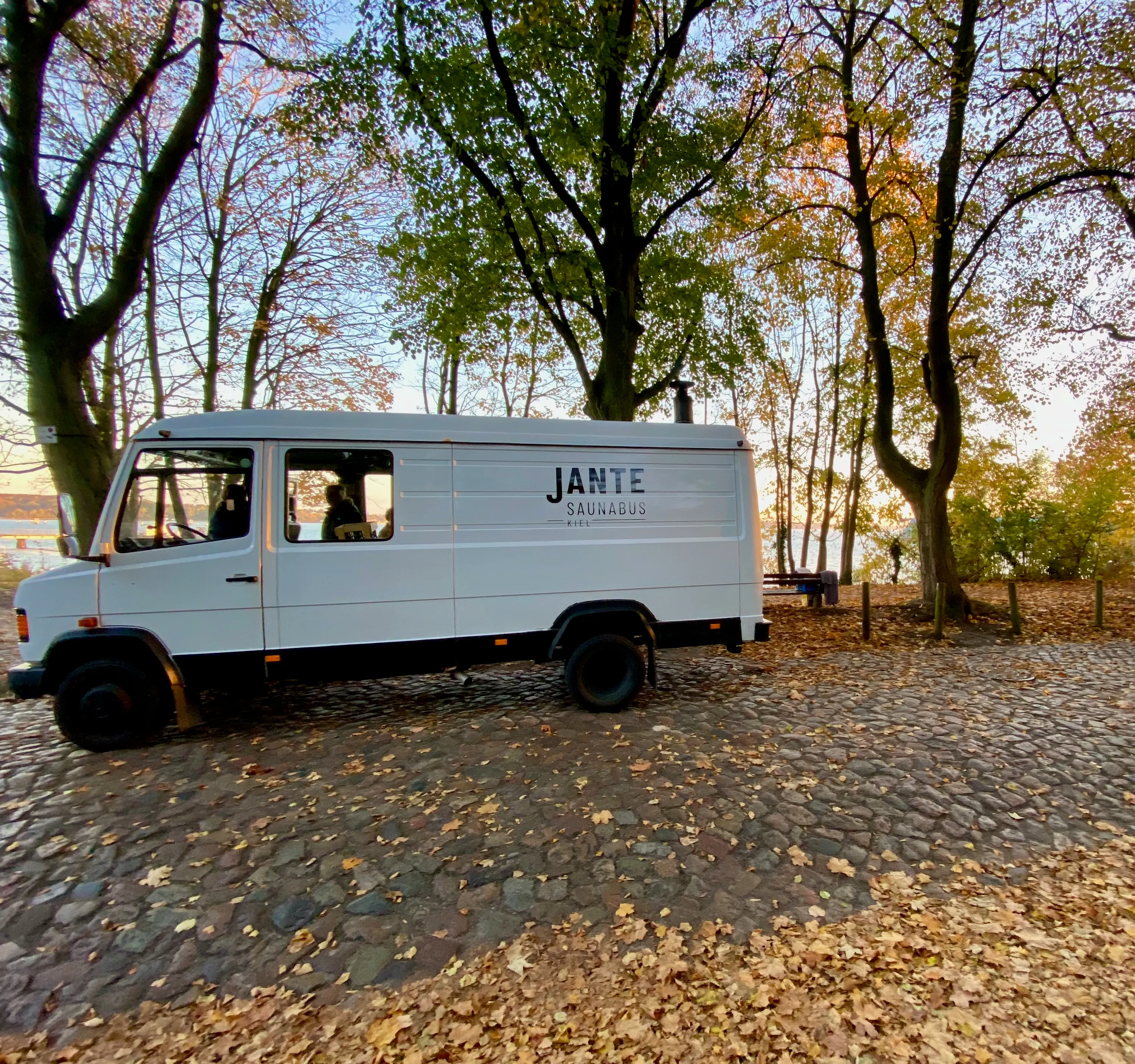 Der Saunabus auf einem mit Herbstlaub bedeckten Kopfsteinpflasterplatz zwischen Bäumen.