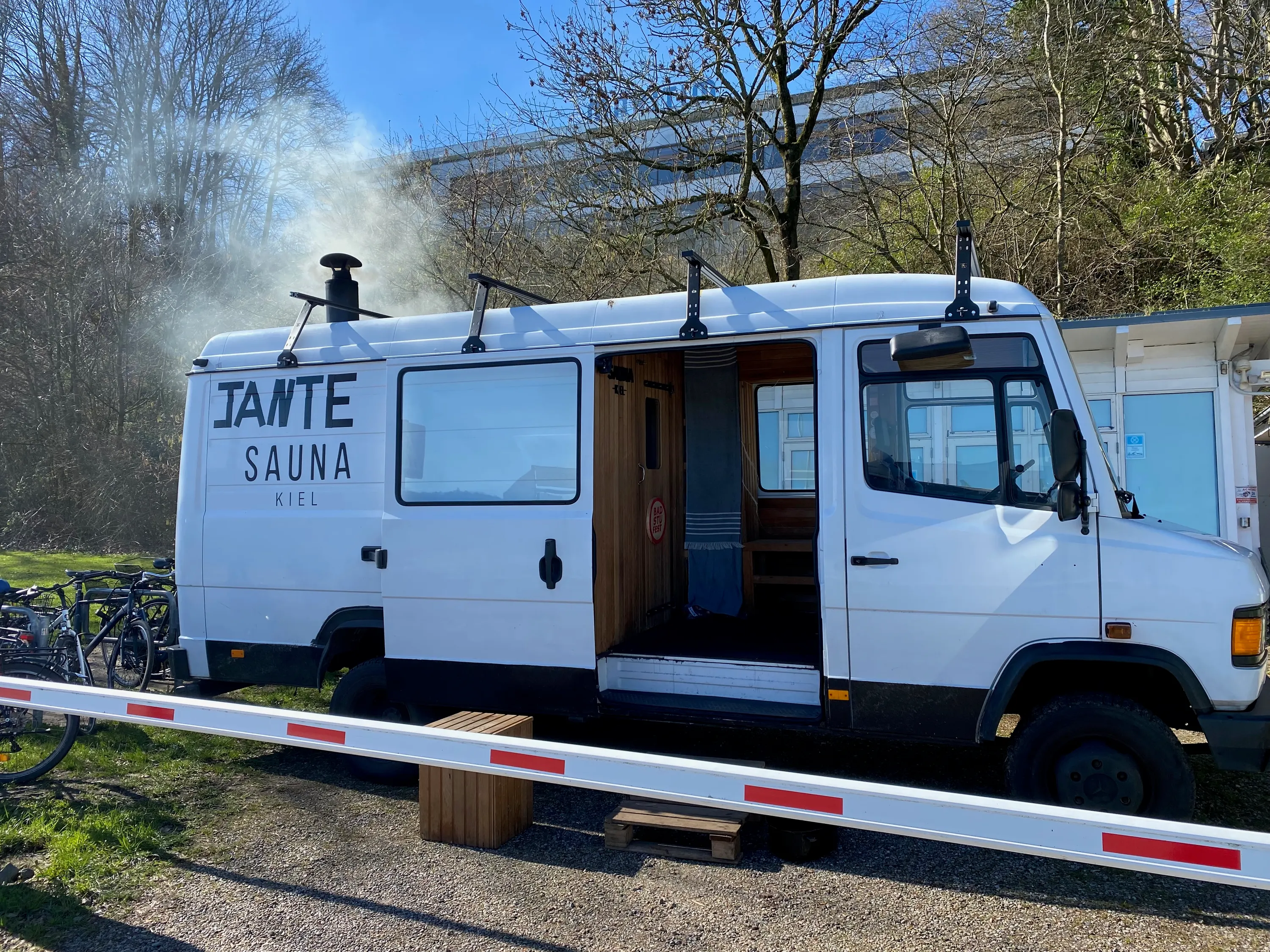 Dunst steigt vom Schornstein des Saunabusses auf, die Tür steht offen, davor liegt eine rot-weiße Schranke.