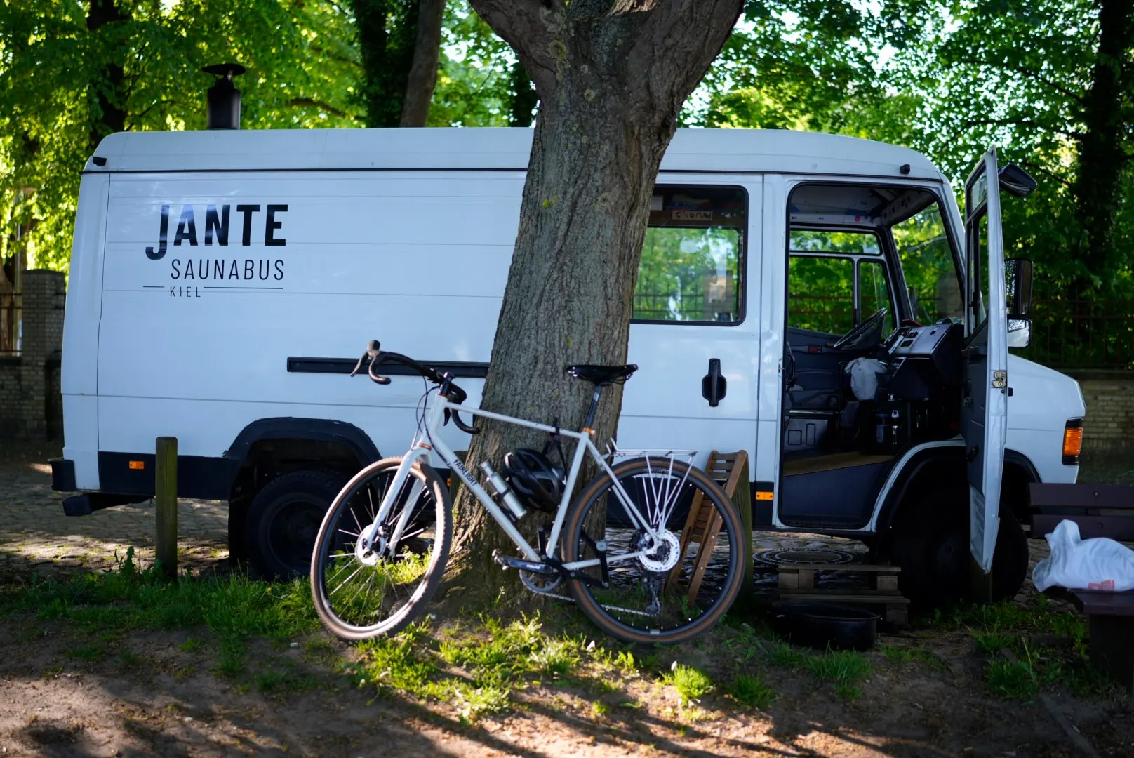 Der Saunabus steht im Schatten eines großen Baums, ein Fahrrad lehnt am Stamm, die Fahrertür steht offen.